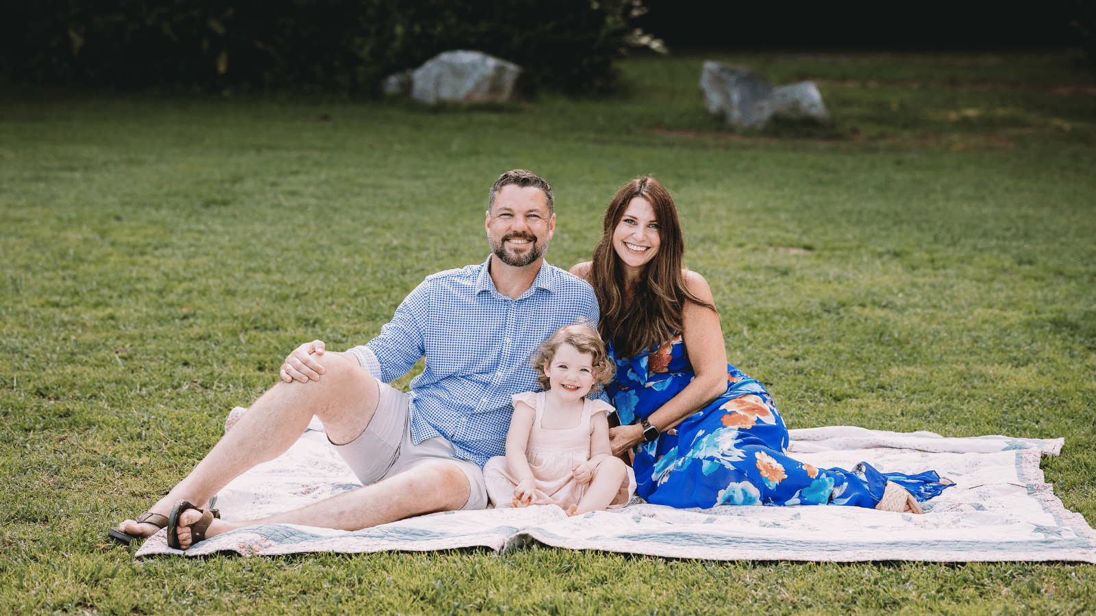 a Cox employee sitting on a blanket in a grass field with her husband and child