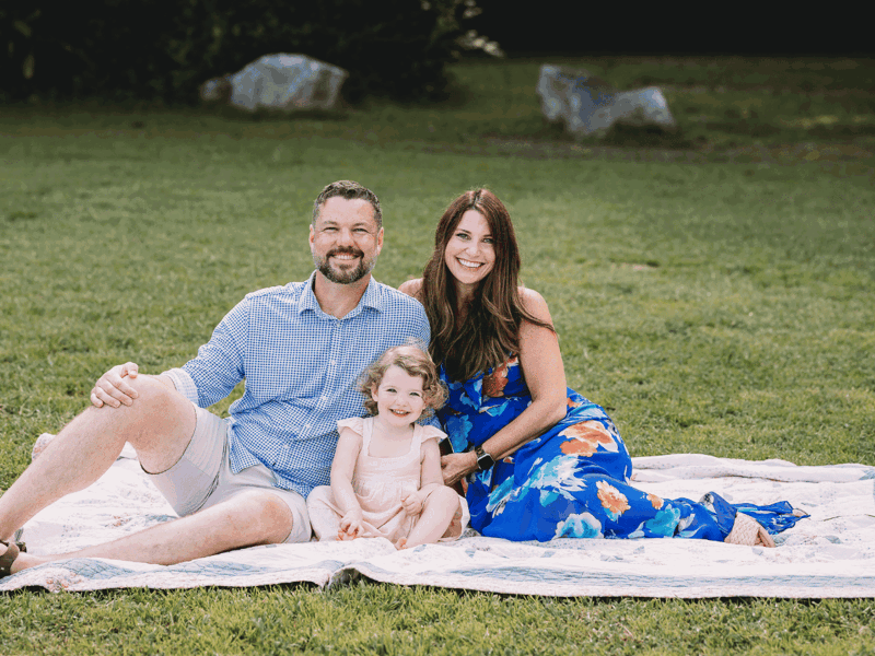 a Cox employee sitting on a blanket in a grass field with her husband and child